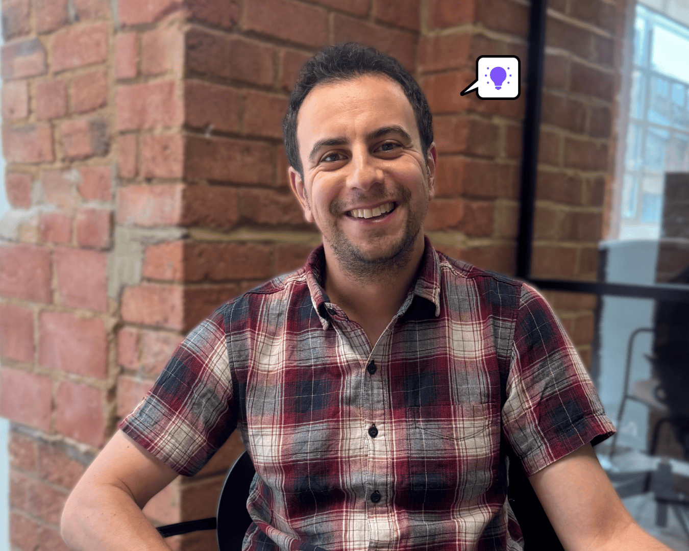 Photo of man with black hair smiling in a checkered shirt with a brick wall background