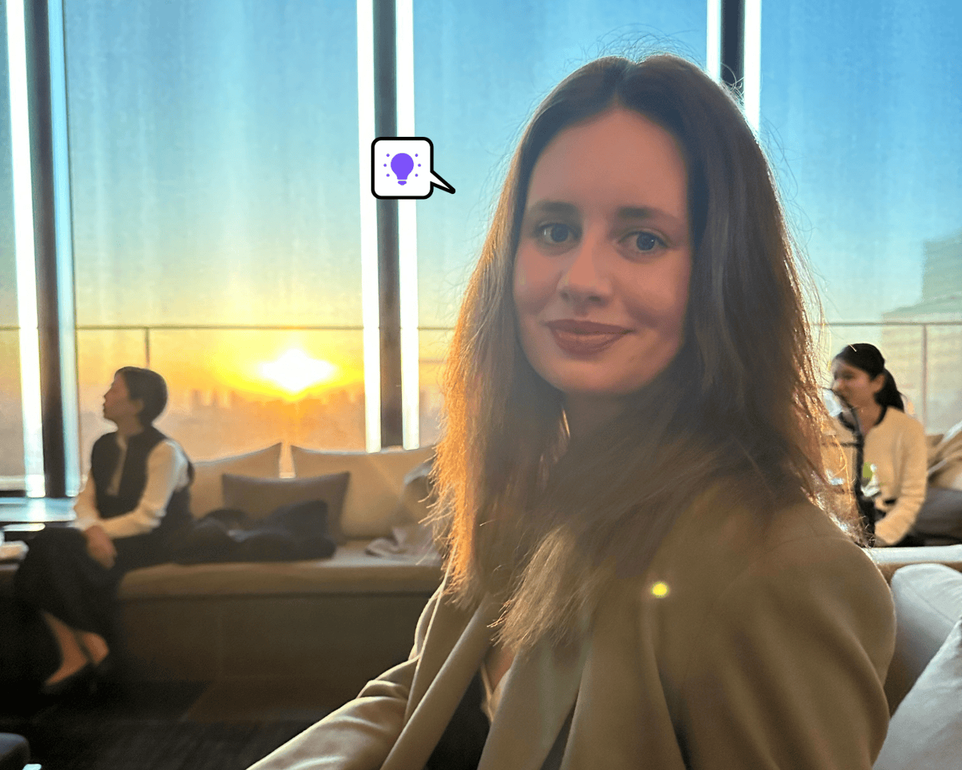 A woman with long brown hair in a blazer smiles at the camera in a modern lounge with large windows. Behind her, the sun is setting over a city skyline, casting a warm glow. Two other women are seated in the background near the window.