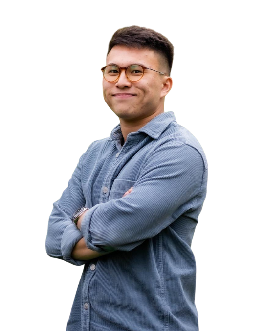 Mid-twenties East Asian man smiles into camera with his arms folded. He's wearing a blue textured button up shirt, and glasses with an orange rim. He has dark hard cropped in short back and sides.
