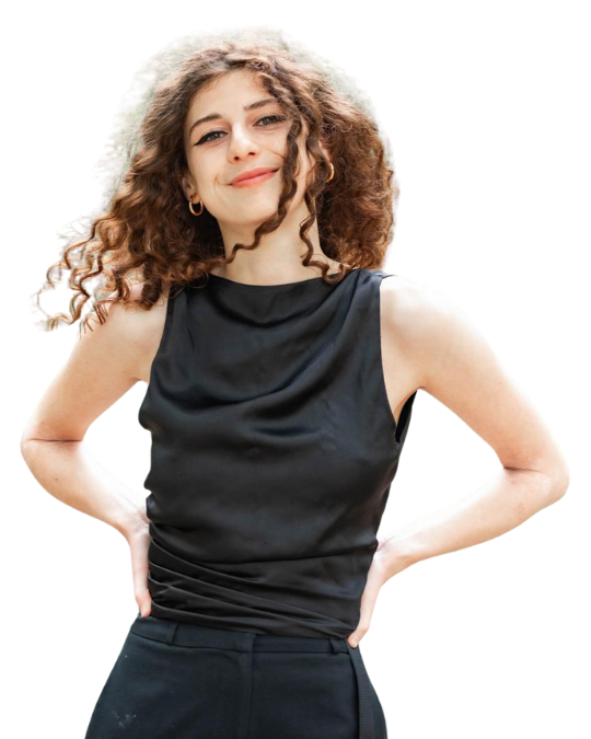 Confident young woman with dark curly hair looks into camera with a bold smile. She's wearing a black high neck satin top, with her hands on both hips