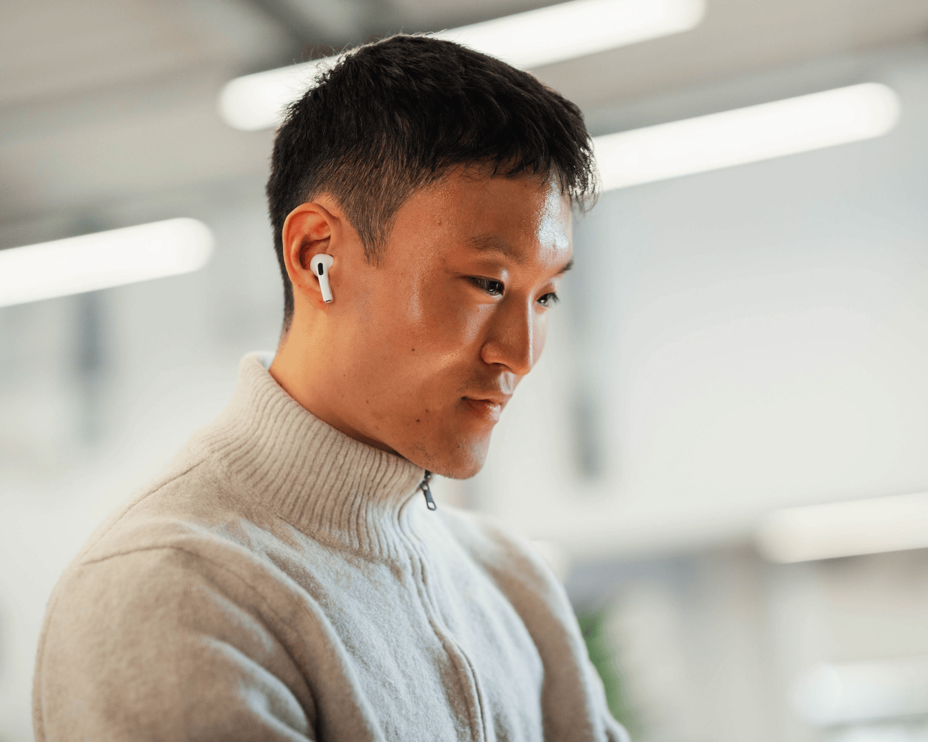 Photo of a man with black hair looking down at a screen with airpods in and wearing a cream zip jumper