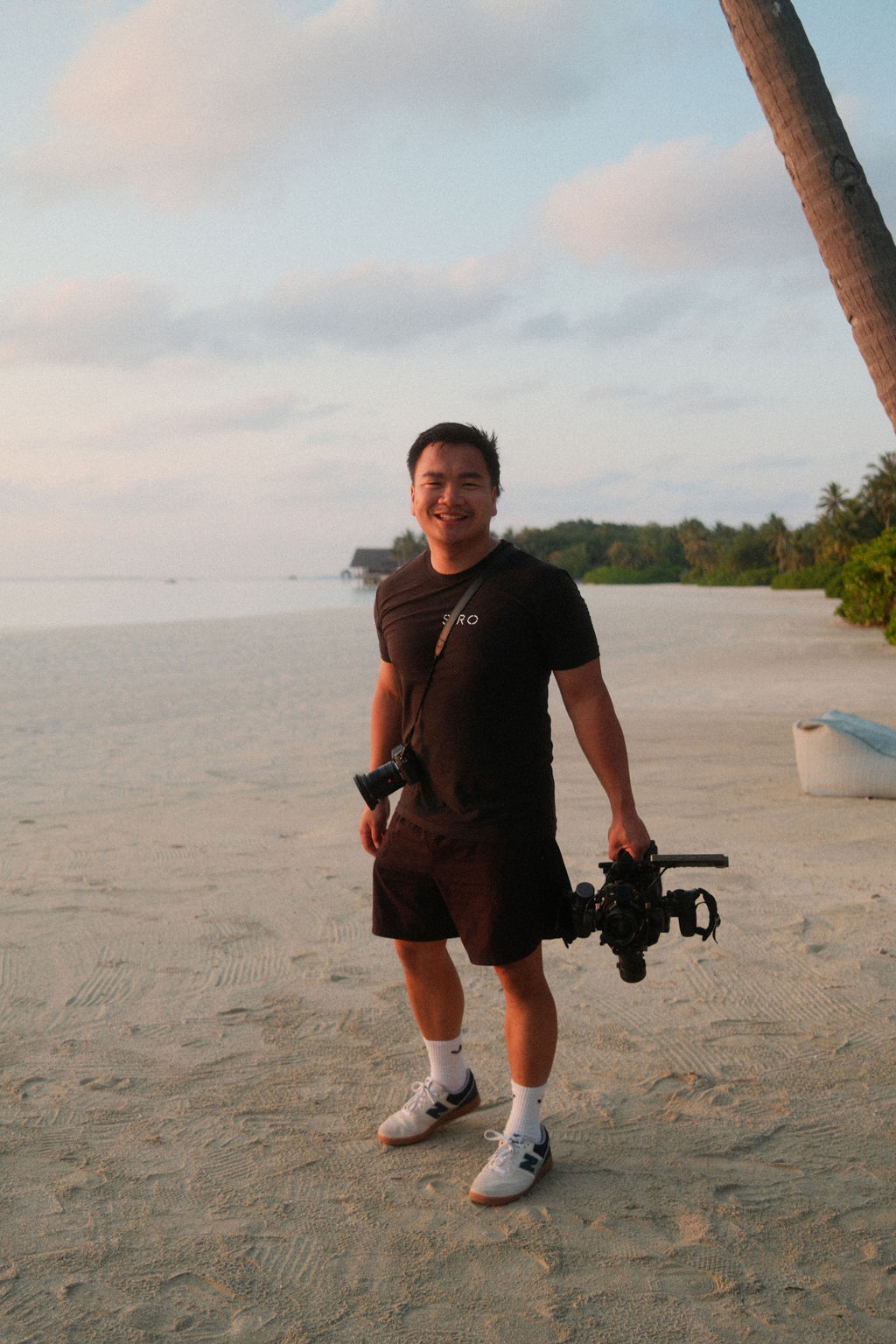 A smiling photographer on a tropical shore holding a camera rig, framed by the sea, palm trees and soft sunset light — evoking wanderlust, creativity and immersive storytelling through authentic imagery.