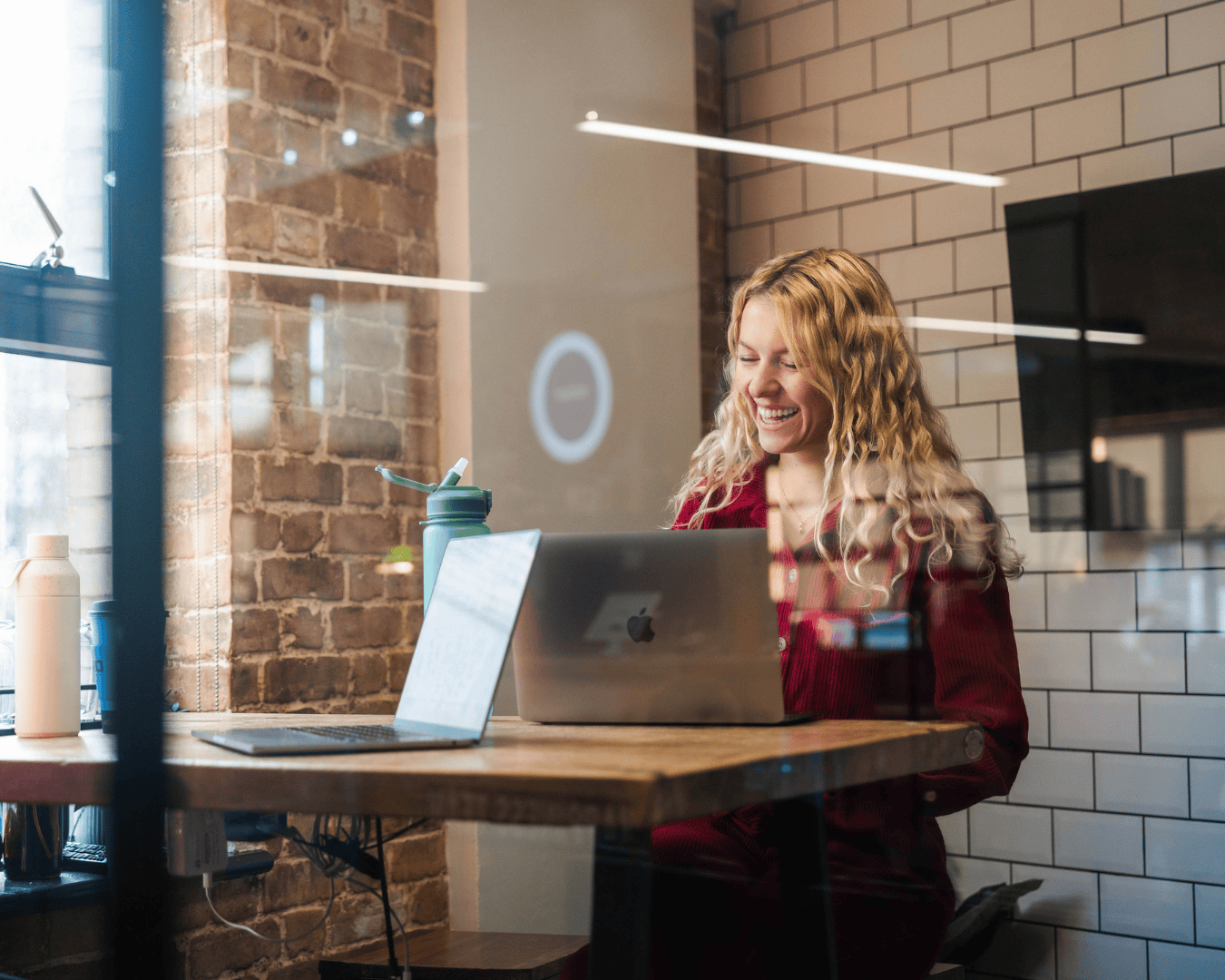 Photo of blonde female smiling at a laptop. She is wearing a red top and in a meeting room