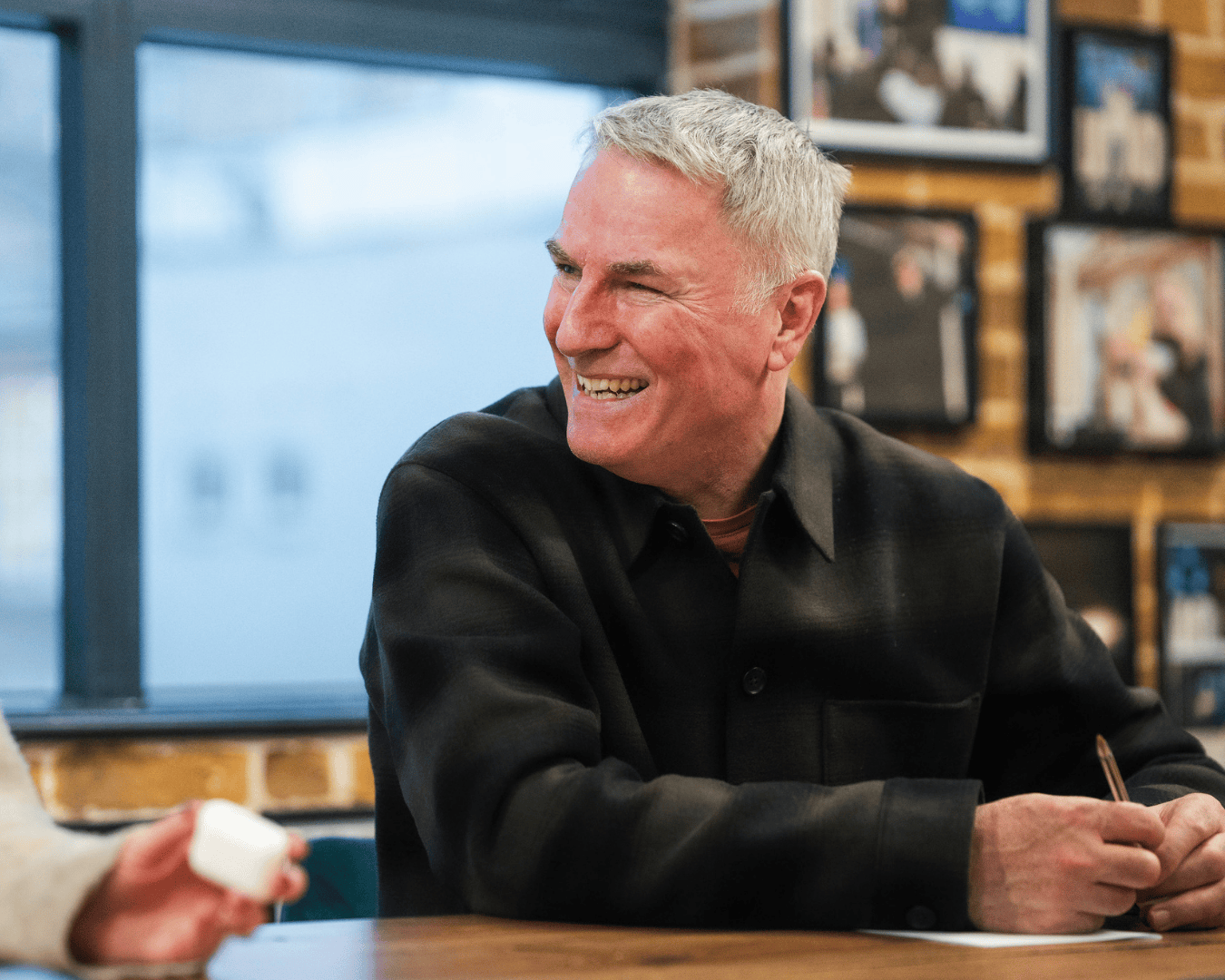man with grey hair smiles warmly while seated at a wooden table in a modern office space, surrounded by brick walls, framed photographs, and a large window behind him.