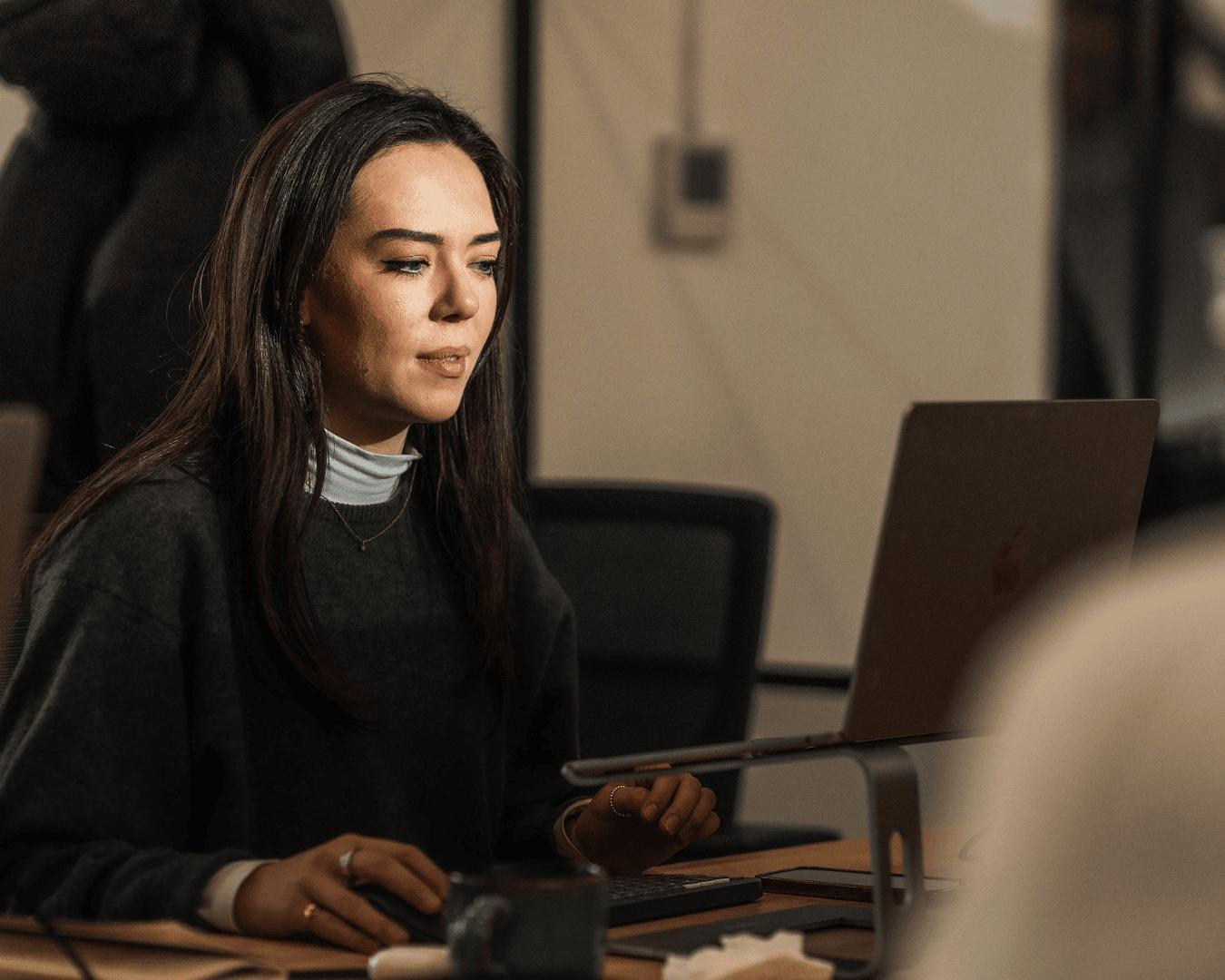 woman with long dark hair works at a laptop on a stand in a dimly lit office, concentrating on her screen, with a mug and paper visible on the desk.
