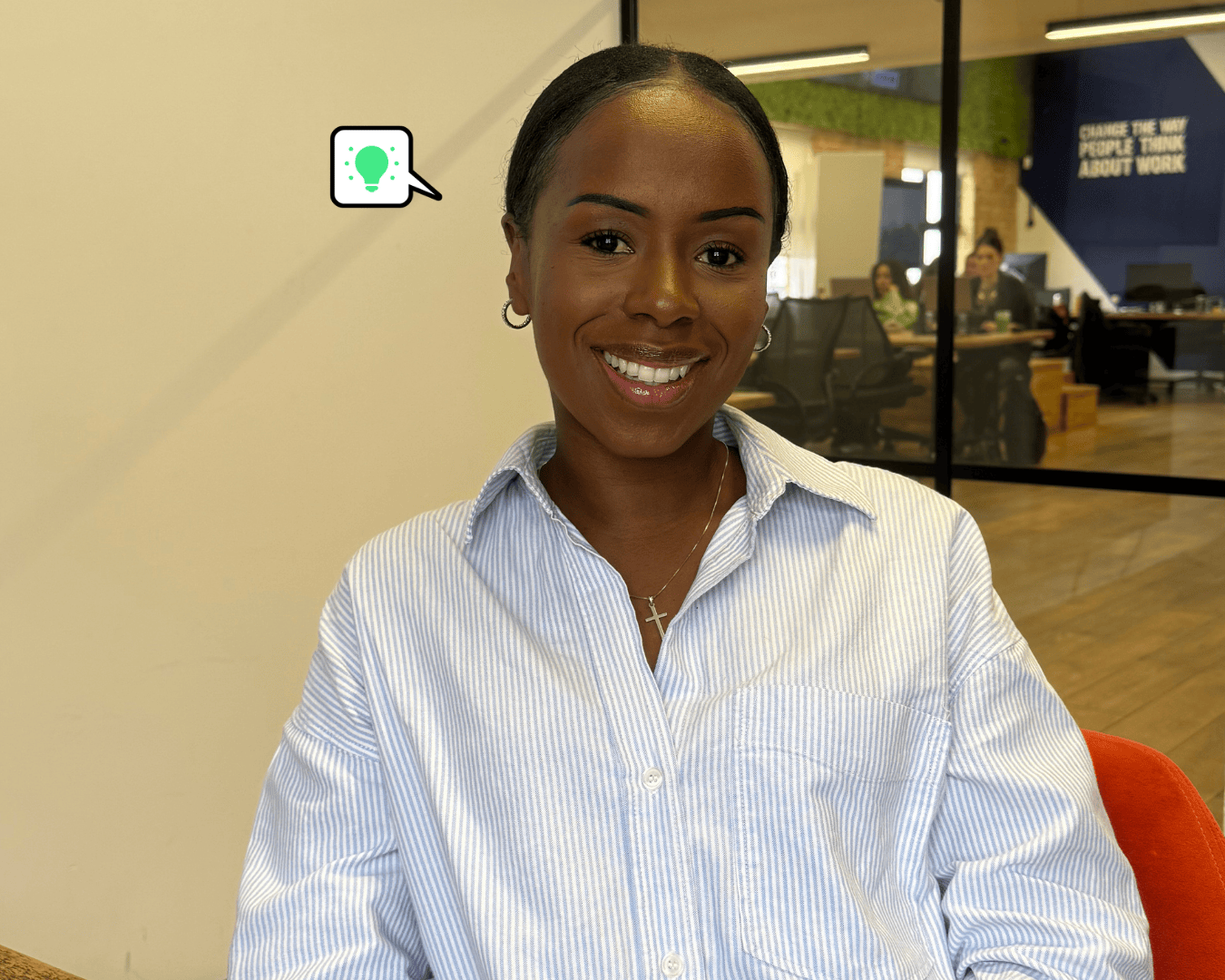Smiling woman in a blue striped shirt sitting in a bright, modern office with colleagues working in the background.