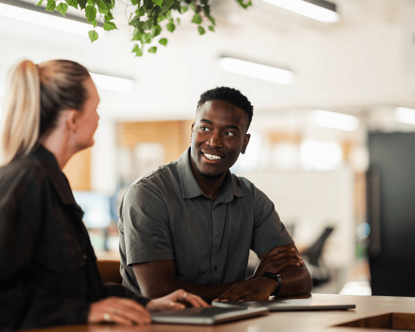 A man and a woman are seated at a table in a brightly lit office, smiling during a conversation. Green plants hang from the ceiling, creating a warm atmosphere.