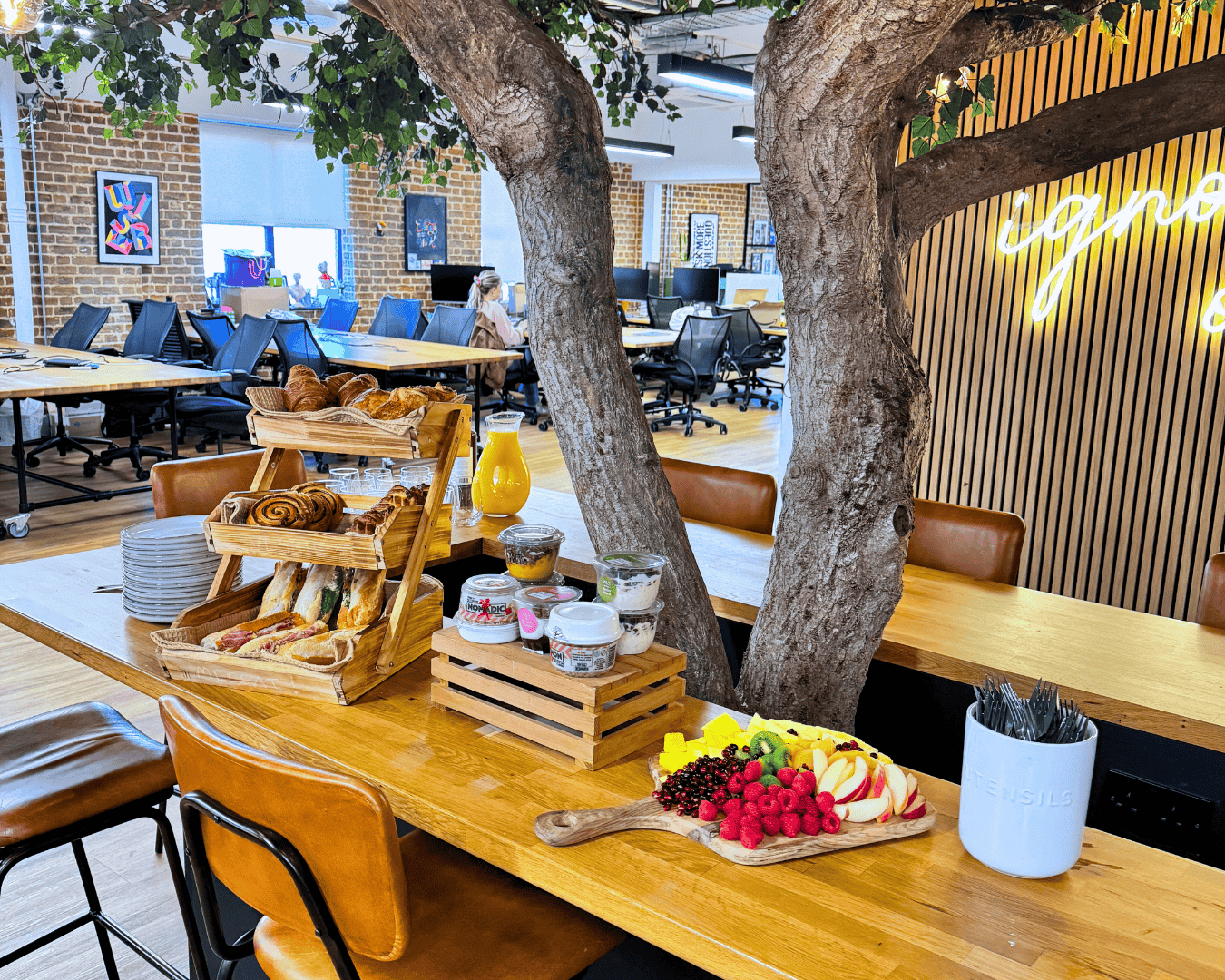 Modern office with a tree centerpiece. A cozy wooden table holds pastries, fruit, and jars accentuated by colorful decor, evoking a vibrant and inviting atmosphere.
