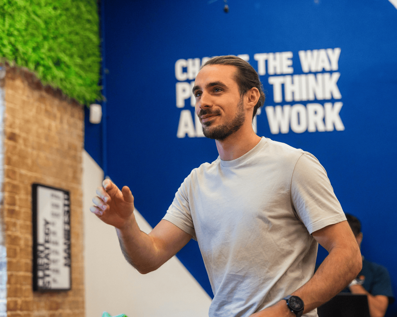 A man in a white t-shirt smiles confidently, gesturing with his hand. Background features motivational text on a blue wall. Bright, positive atmosphere.