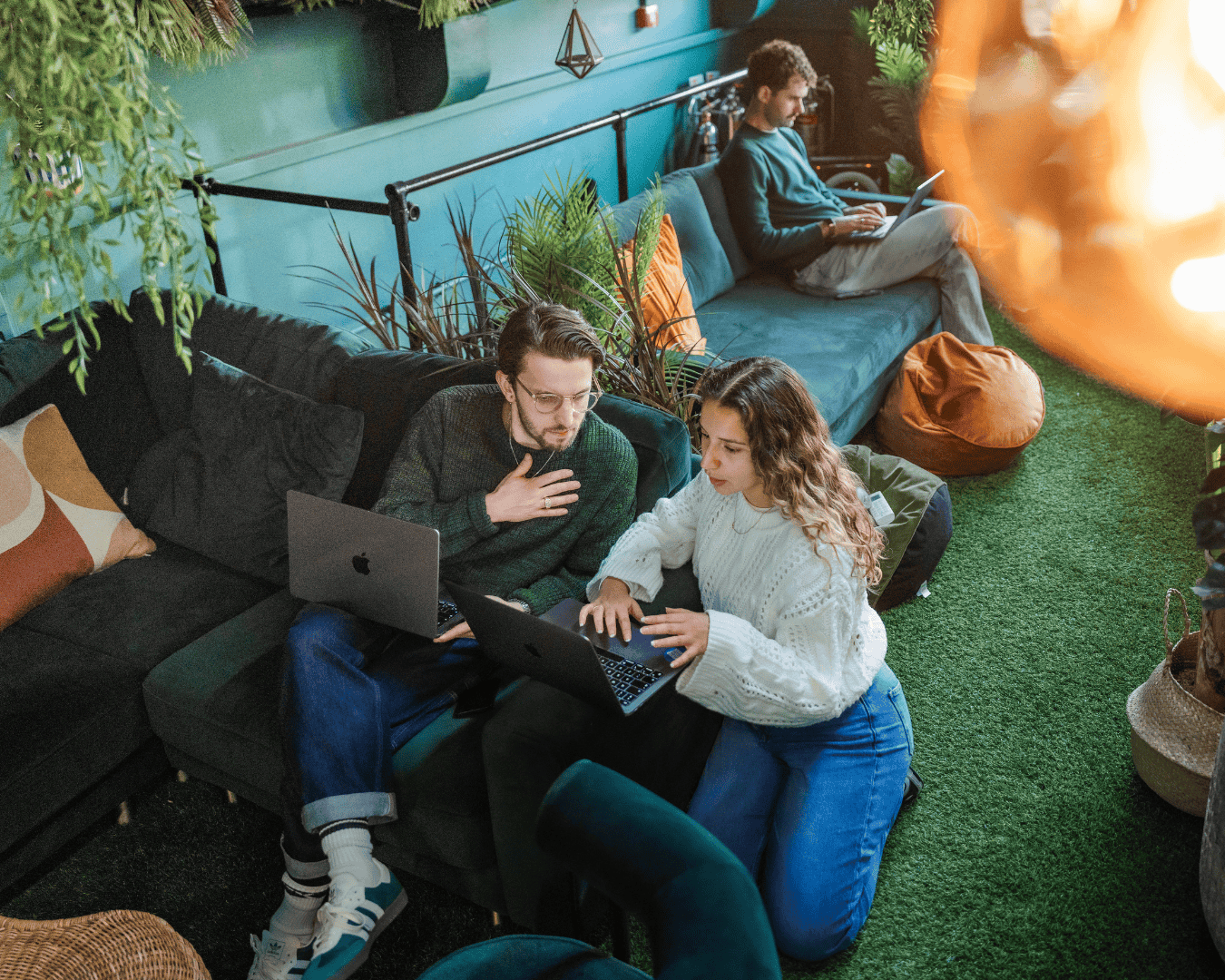 Two people collaborate on a laptop in a cozy, green indoor setting with plants and soft seating. A third person works on another laptop in the background.