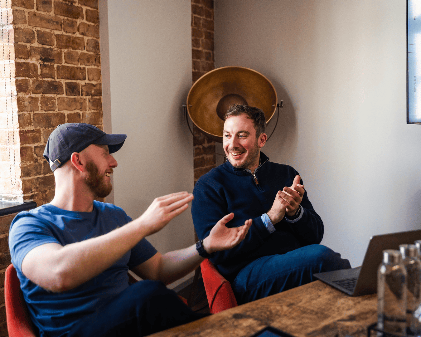 Two men sit in a brick-walled office, smiling and gesturing energetically. A laptop is on the table, and a presentation is displayed on a screen.