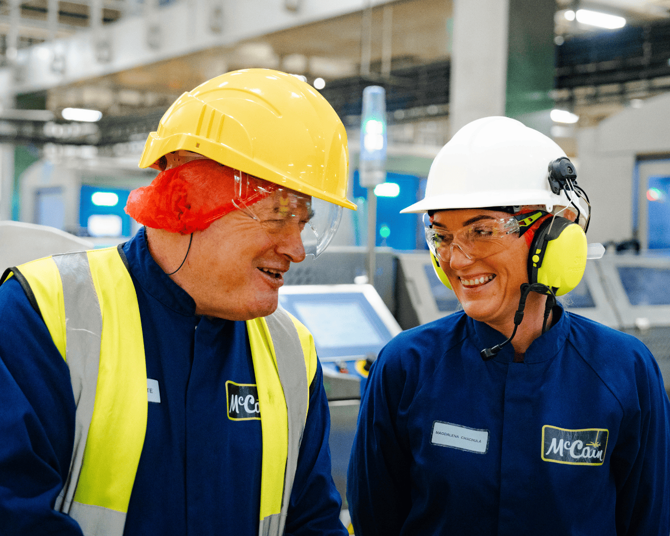 Two factory workers in blue uniforms and safety gear share a lighthearted moment. They wear hard hats and protective glasses, smiling warmly. Industrial setting.
