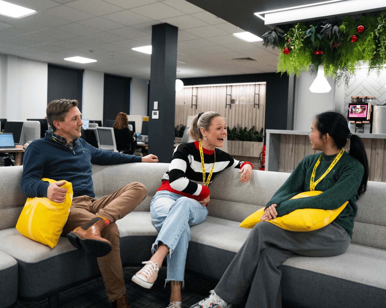 A group of three colleagues sits on a gray couch in an office lounge, smiling and laughing. They hold yellow pillows, and greenery hangs overhead. Warm, relaxed atmosphere.