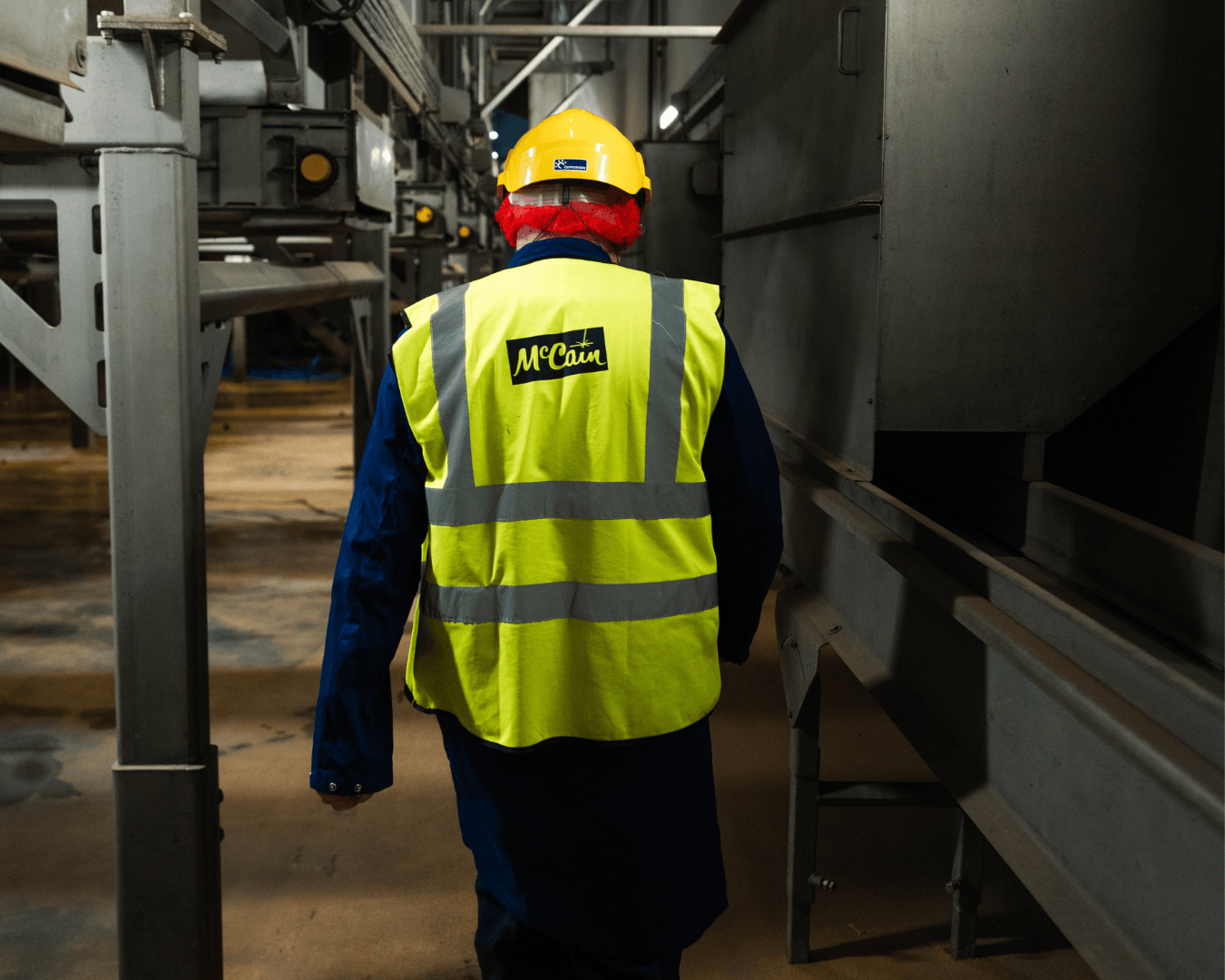 A worker wearing a yellow hard hat and high-visibility vest labeled "McCain" walks through an industrial setting, conveying focus and safety.