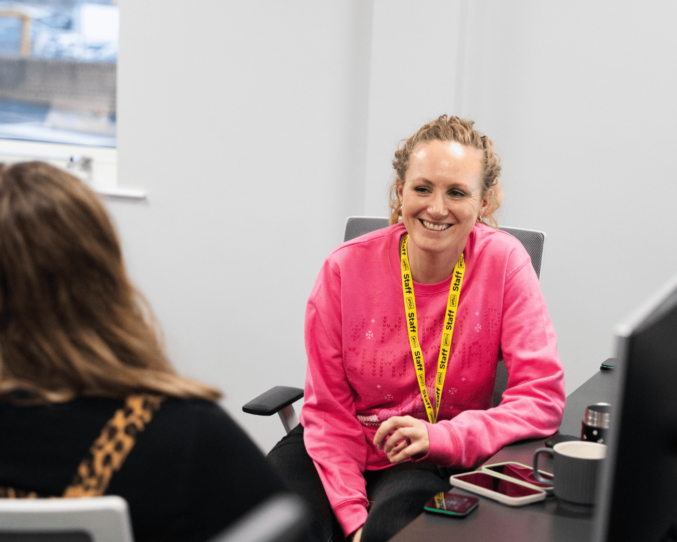 A woman in a bright pink sweater with a yellow lanyard smiles warmly while sitting at a desk, engaging in a conversation with another person.