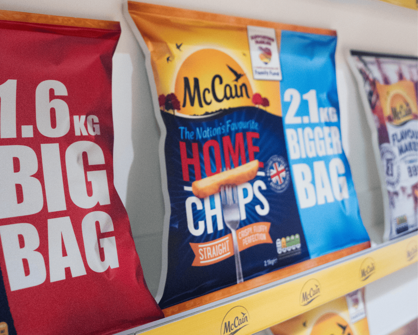 Bags of McCain chips on a shelf. A red bag reads "1.6 kg BIG BAG" and a blue one reads "2.1 kg BIGGER BAG." Central bag showcases "Home Chips."