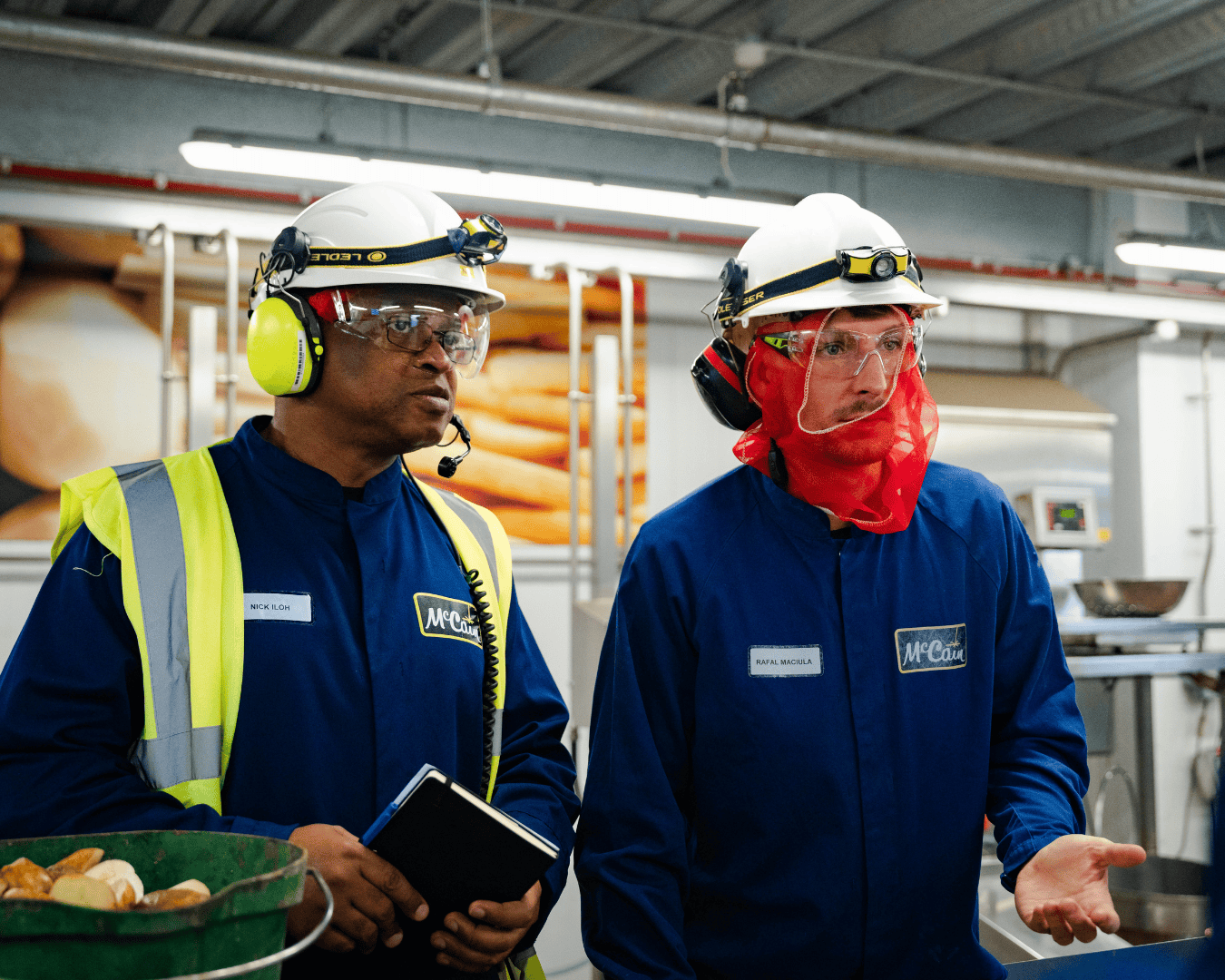 Two workers in blue coveralls and safety gear stand in an industrial setting. One holds a notebook; both appear focused and engaged in discussion.