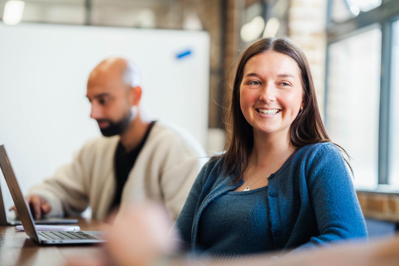 Smiling woman in a blue cardigan sits at a table, facing camera. A colleague works on a laptop in the soft-focus background. Bright windows and brick walls suggest a workspace.