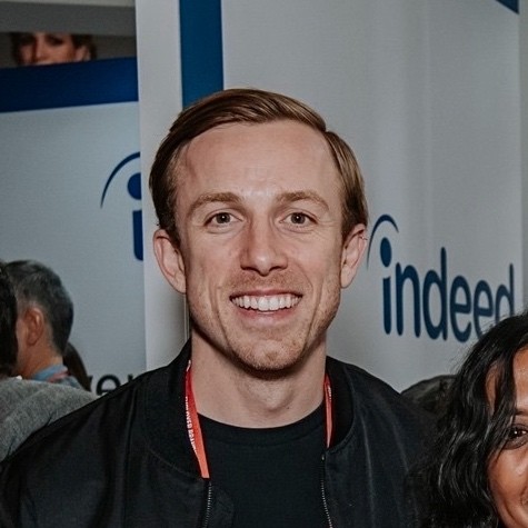 Smiling man with short light-brown hair poses indoors at an event, wearing a black jacket and red lanyard. Branded Indeed signage and attendees appear in the background of the booth.