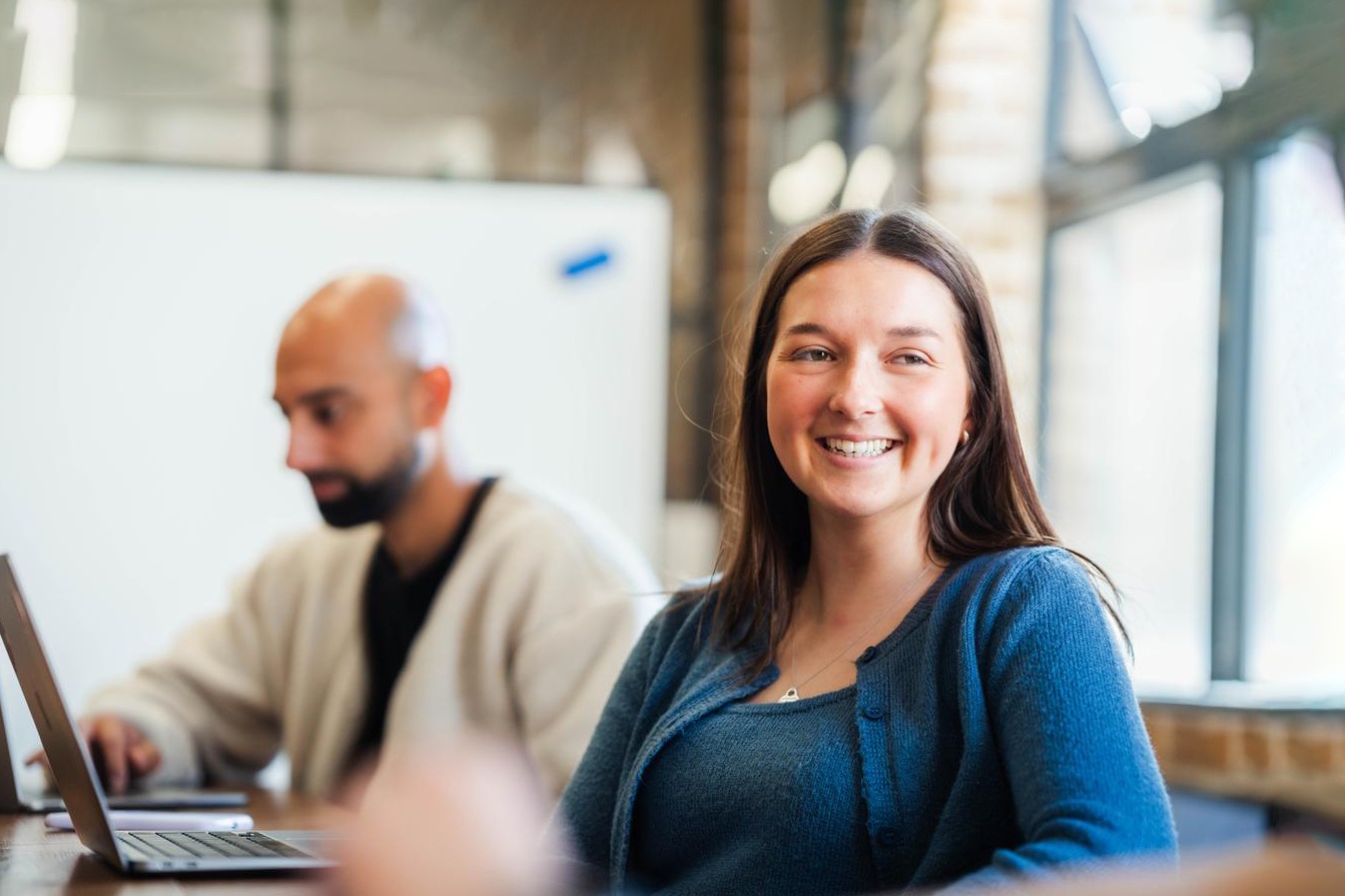Smiling woman in a blue cardigan sits at a table, facing camera. A colleague works on a laptop in the soft-focus background. Bright windows and brick walls suggest a workspace.