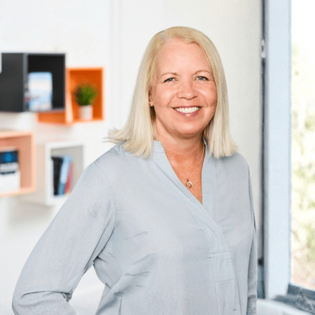 A smiling woman with straight blonde hair wearing a light grey blouse, standing confidently in a modern office space with geometric shelving and a window letting in natural light.
