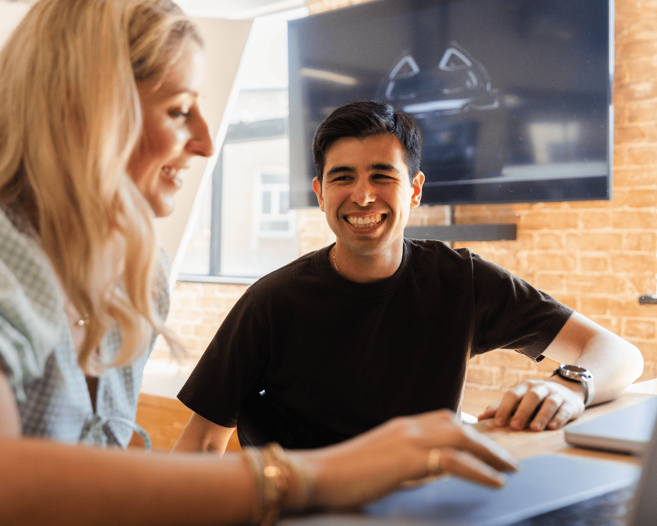 A man and a woman are smiling and engaging in conversation at a desk. A large screen is visible in the background, creating a warm, collaborative atmosphere.