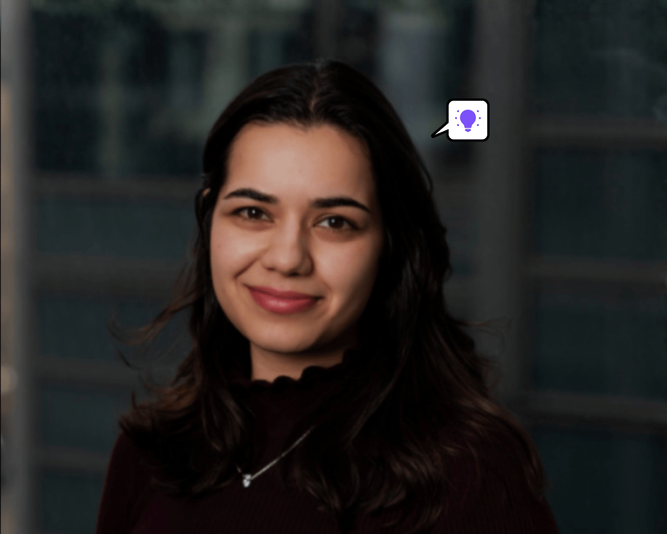 Smiling woman with dark hair stands indoors, wearing a dark top. A speech bubble with a light bulb icon hovers beside her, suggesting an idea.