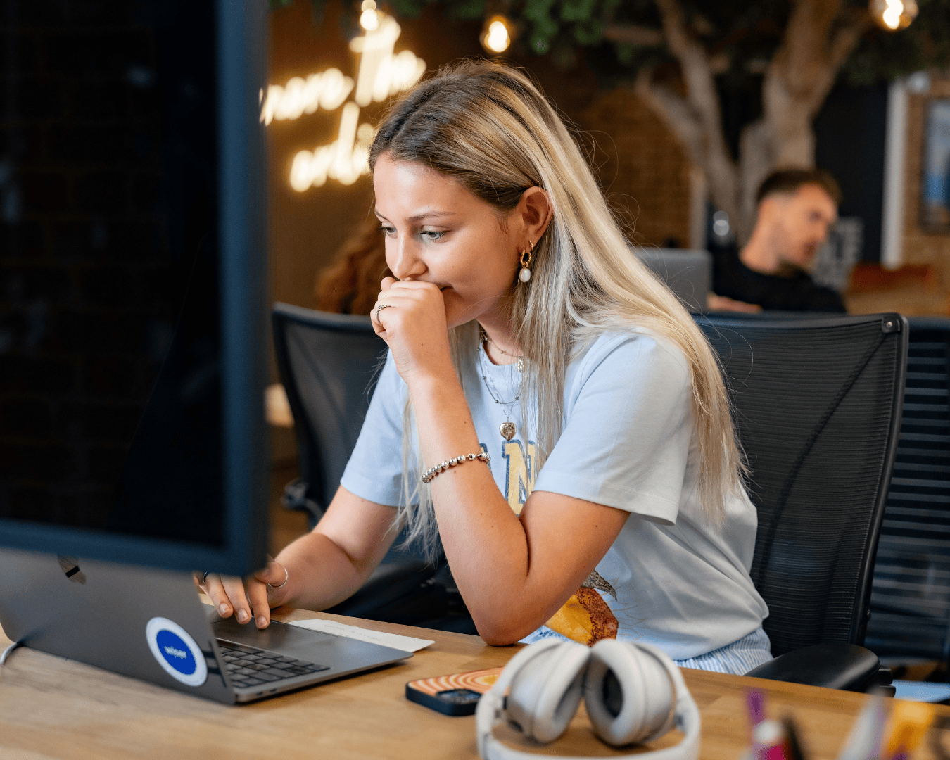 A woman with long blonde hair focuses intently on a laptop at a modern desk, surrounded by tech gadgets. A blurred coworker is in the background.