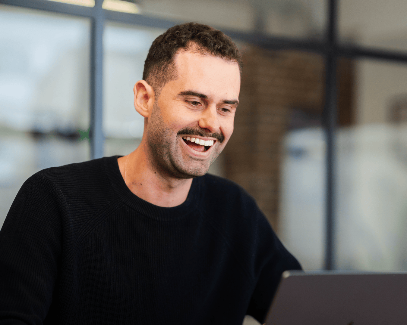 A man with a mustache, wearing a black sweater, smiles while looking at his laptop. The background shows a blurred office setting with a large window.