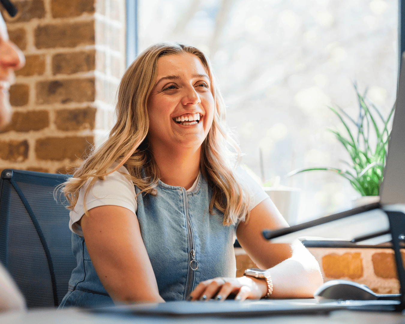 Smiling woman with long blonde hair, wearing a denim top, sits in a bright office with a brick wall. A plant and open laptop are in view. Warm, cheerful mood.