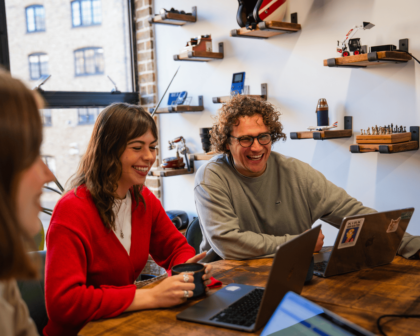 A group of three people, two women and a man, sit around a table with laptops, smiling and laughing. The room has shelves with models and decor.