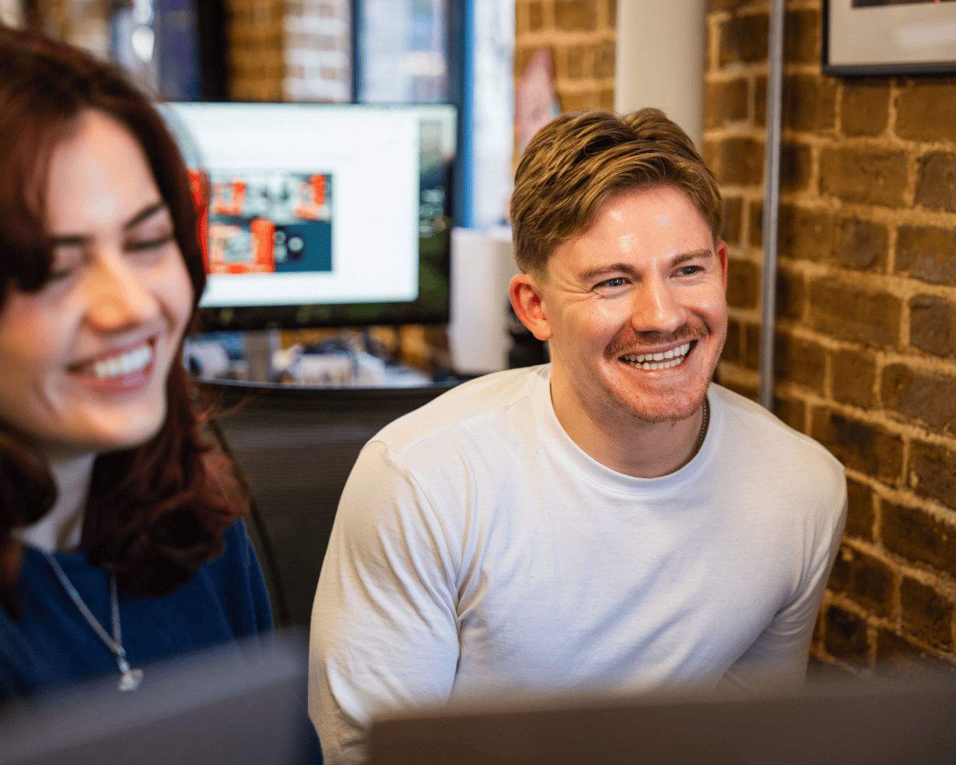 Two people smiling in an office setting with exposed brick walls. A computer displays a blurred screen in the background, creating a cheerful atmosphere.