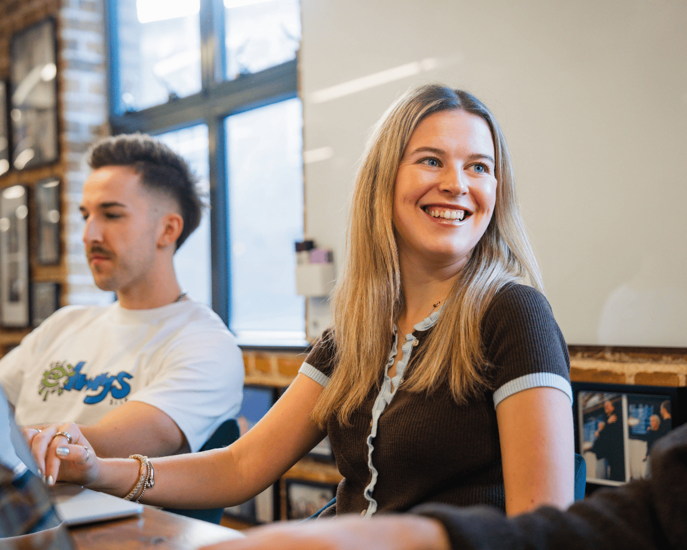 A woman smiling during a meeting in a bright office with brick walls and framed photos. A man sits beside her, focused on a laptop. Warm, collaborative atmosphere.