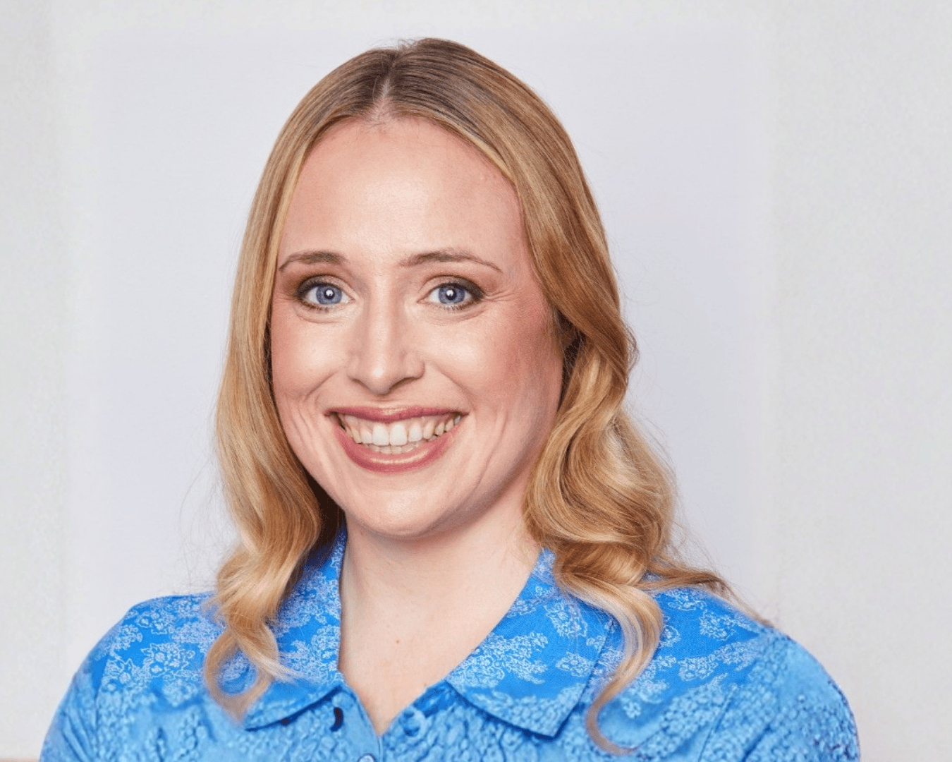 A woman with long blonde hair smiles warmly, wearing a bright blue patterned blouse. The background is plain white.