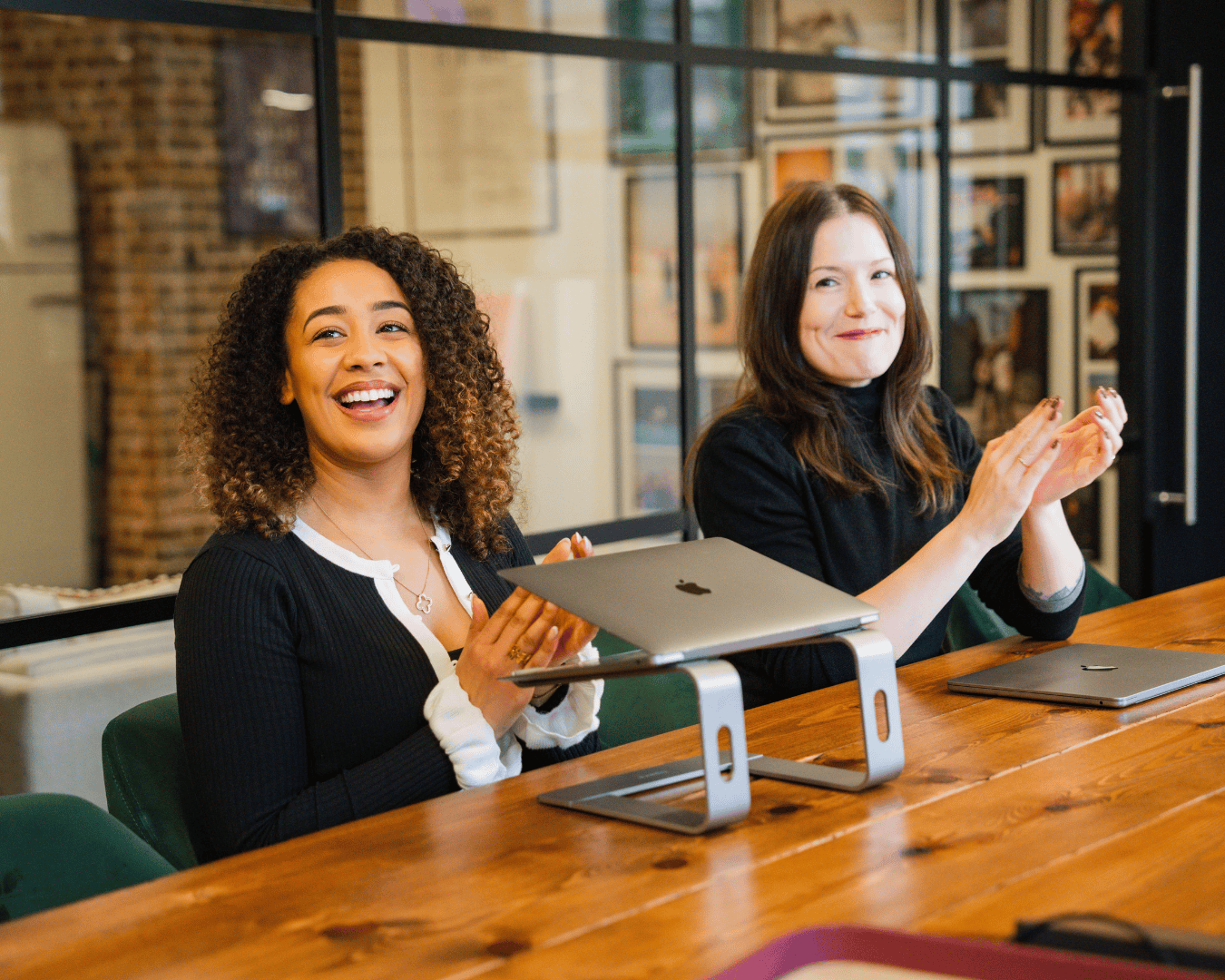 Two women sit at a wooden table with laptops, smiling and clapping. The setting appears to be a modern, well-lit office, conveying a cheerful and collaborative atmosphere.
