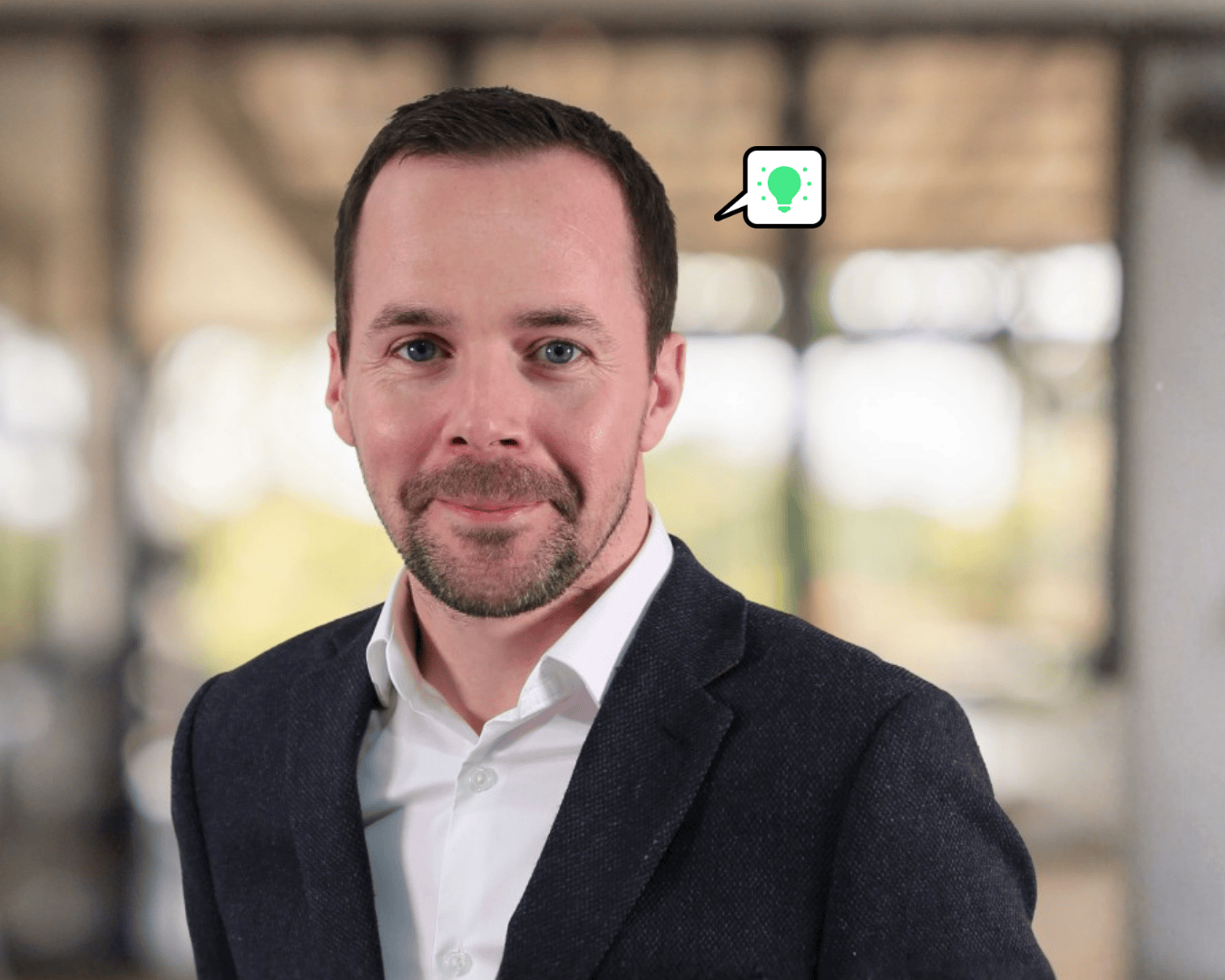 A smiling man in a suit, standing indoors with a blurred background. A speech bubble with a green light bulb icon is near his head, suggesting an idea.