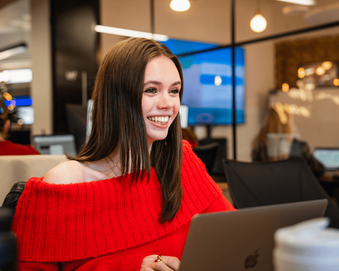 Smiling woman in a bright red sweater works on a laptop in a modern office. She appears focused and happy, with a large screen behind her.