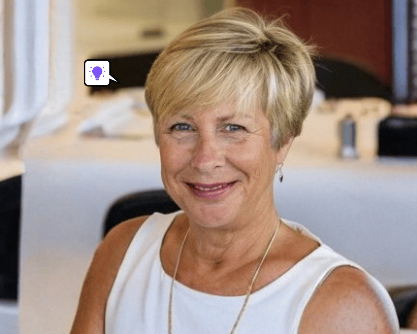 Smiling woman with short blonde hair in a white sleeveless top, seated indoors. A speech bubble with a purple lightbulb icon appears nearby.