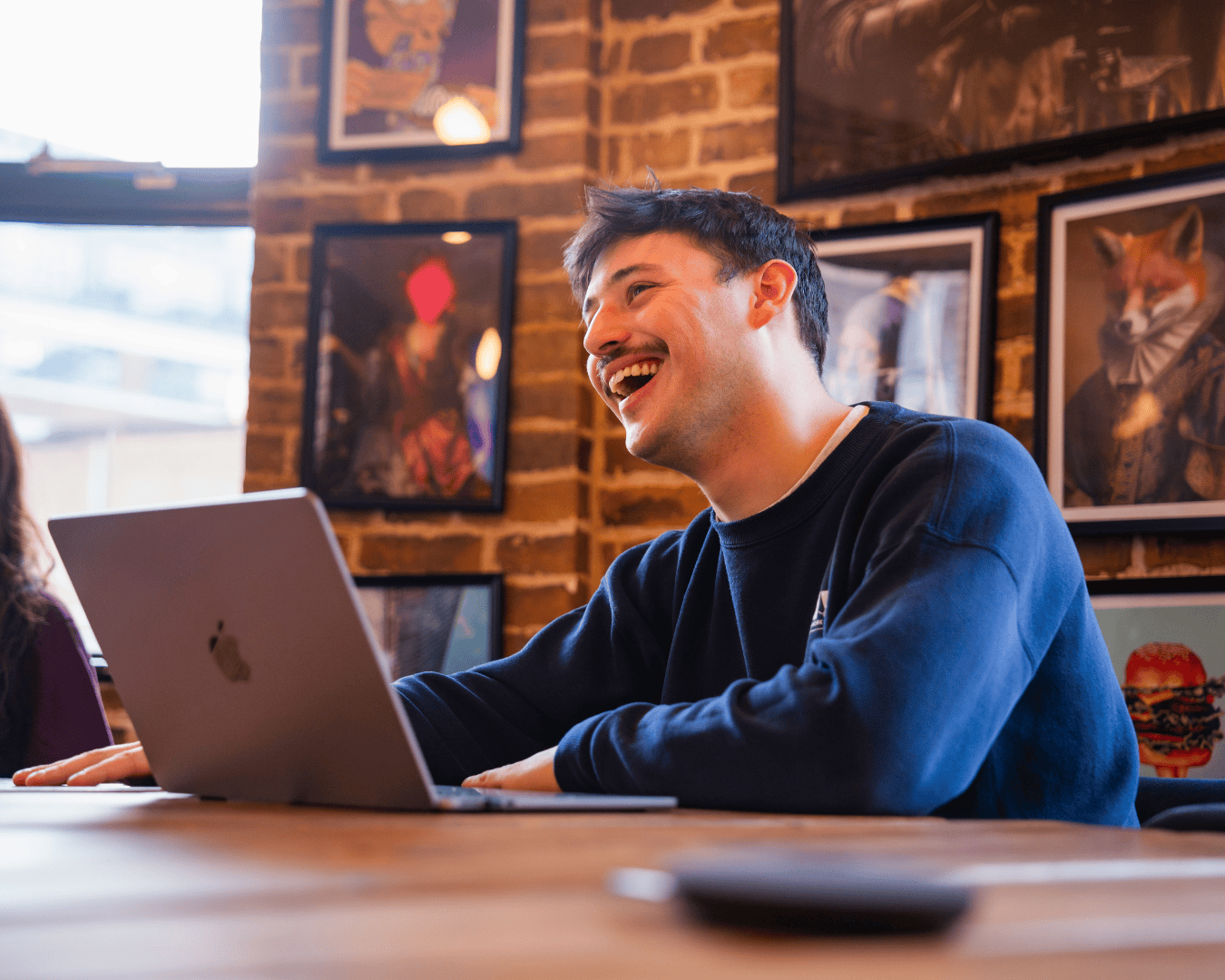 A man with a mustache is happily talking, seated at a table with a laptop. A woman sits nearby. Brightly lit brick room, framed art on walls