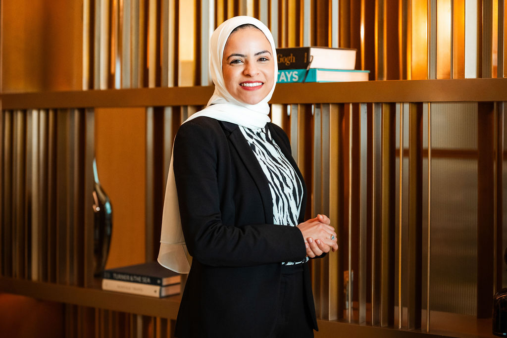 Professional woman wearing a headscarf and business attire stands smiling in a modern interior, with shelves, books, and warm lighting creating a polished, welcoming atmosphere.