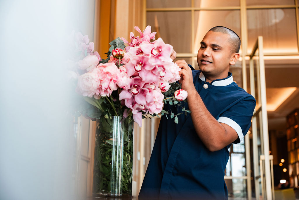 Staff member carefully arranges a tall floral display in a hotel interior, adjusting pink flowers in a glass vase within a softly lit, refined hospitality environment.