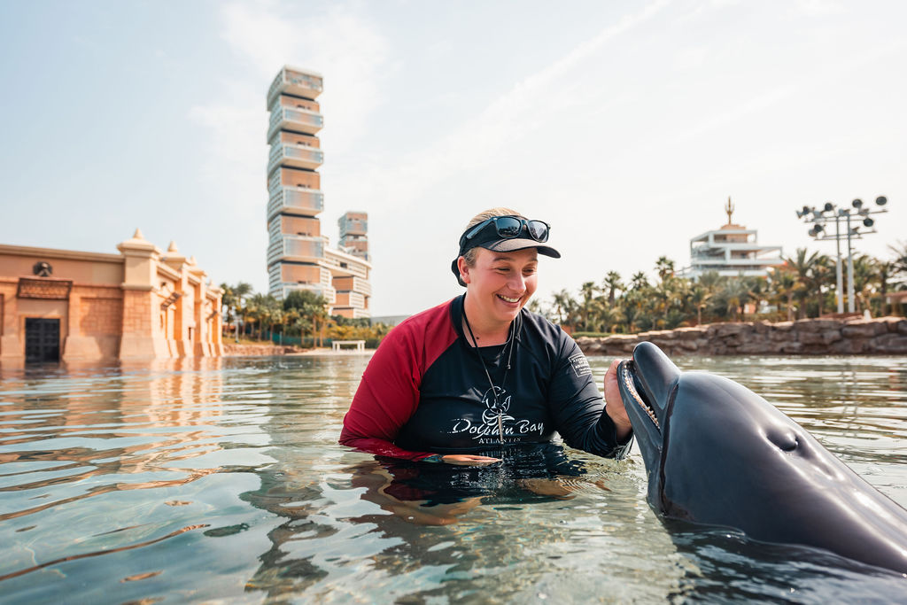 Adult woman in her 30s with light brown hair tucked under a cap smiles while standing waist-deep in water, gently interacting with a dolphin at an outdoor marine habitat.