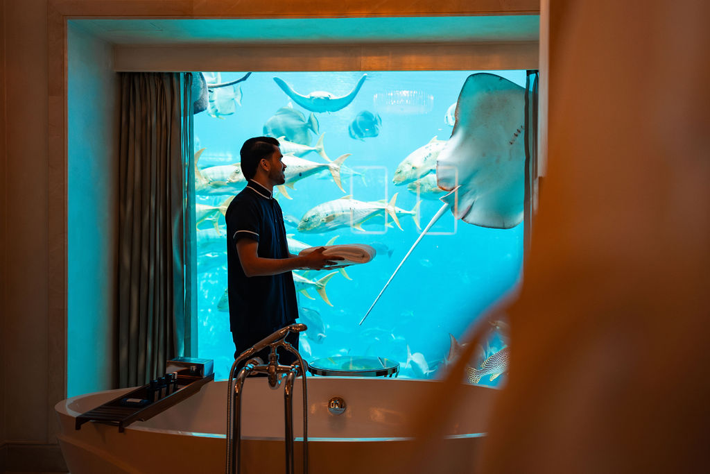 Adult man in his 20s or 30s with short dark hair stands beside a freestanding bath, holding folded towels, with large fish swimming past an aquarium window behind him.