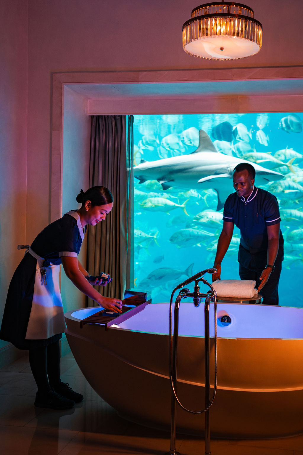 Two adult hospitality staff members, a woman and a man in their 20s or 30s, with dark hair, prepare a freestanding bath in a luxury room with a large aquarium window.