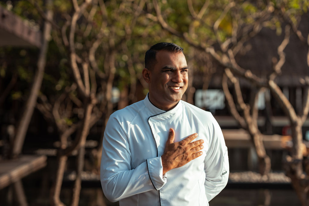 Adult man in his 30s with short dark hair smiles warmly outdoors, wearing a white chef’s jacket and placing his hand over his chest, with trees and soft sunlight behind him.