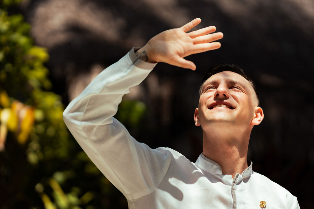 Adult man in his 30s with short dark hair raises his hand to shade his eyes, smiling while standing outdoors in sunlight, wearing a light uniform amid greenery.