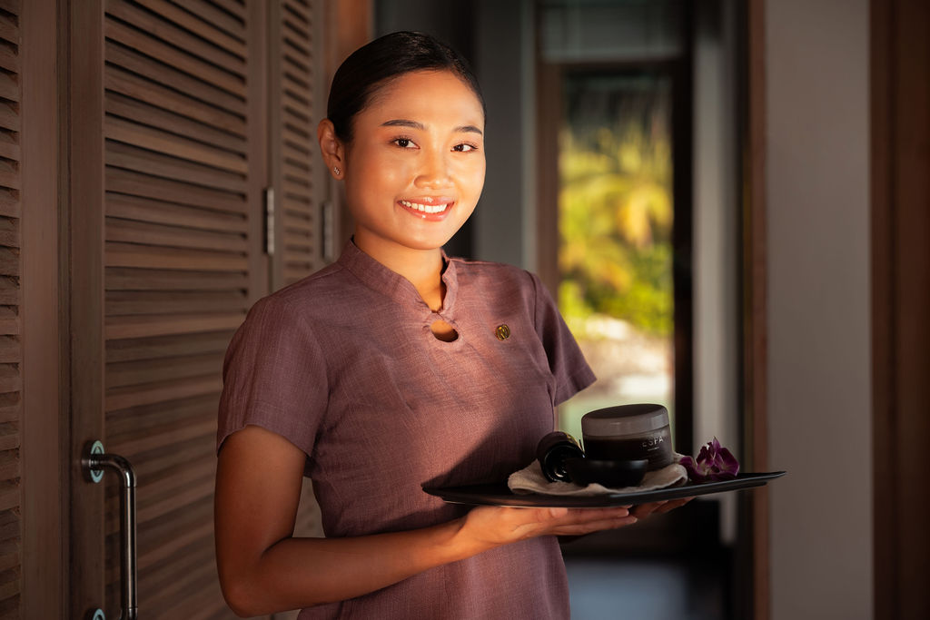 Adult woman in her 20s or 30s with dark hair neatly tied back smiles while holding a tray with spa items, standing in a softly lit doorway of a treatment room.