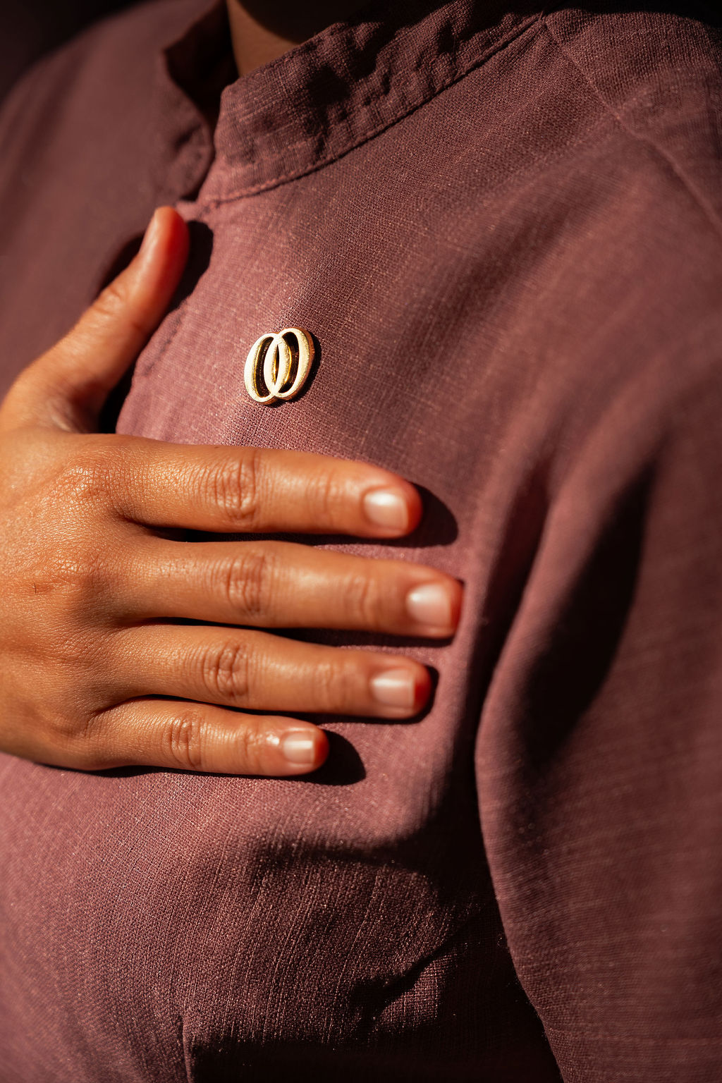 Close-up of adult woman’s hand resting over her chest on a mauve spa uniform, showing a small gold pin, with soft light highlighting fabric texture and gentle gesture.
