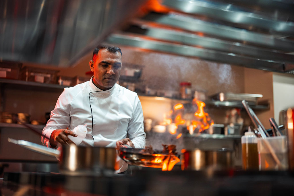 Adult man in his 30s with short dark hair cooks at a professional stove, wearing a white chef’s jacket while stirring a pan in a busy, warmly lit kitchen.