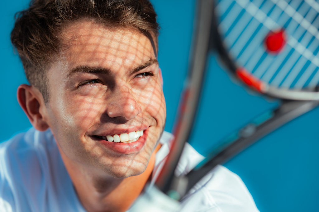 Adult man in his 20s or 30s with short light brown hair smiles while holding a tennis racket, sunlight casting patterned shadows across his face against a bright blue background.